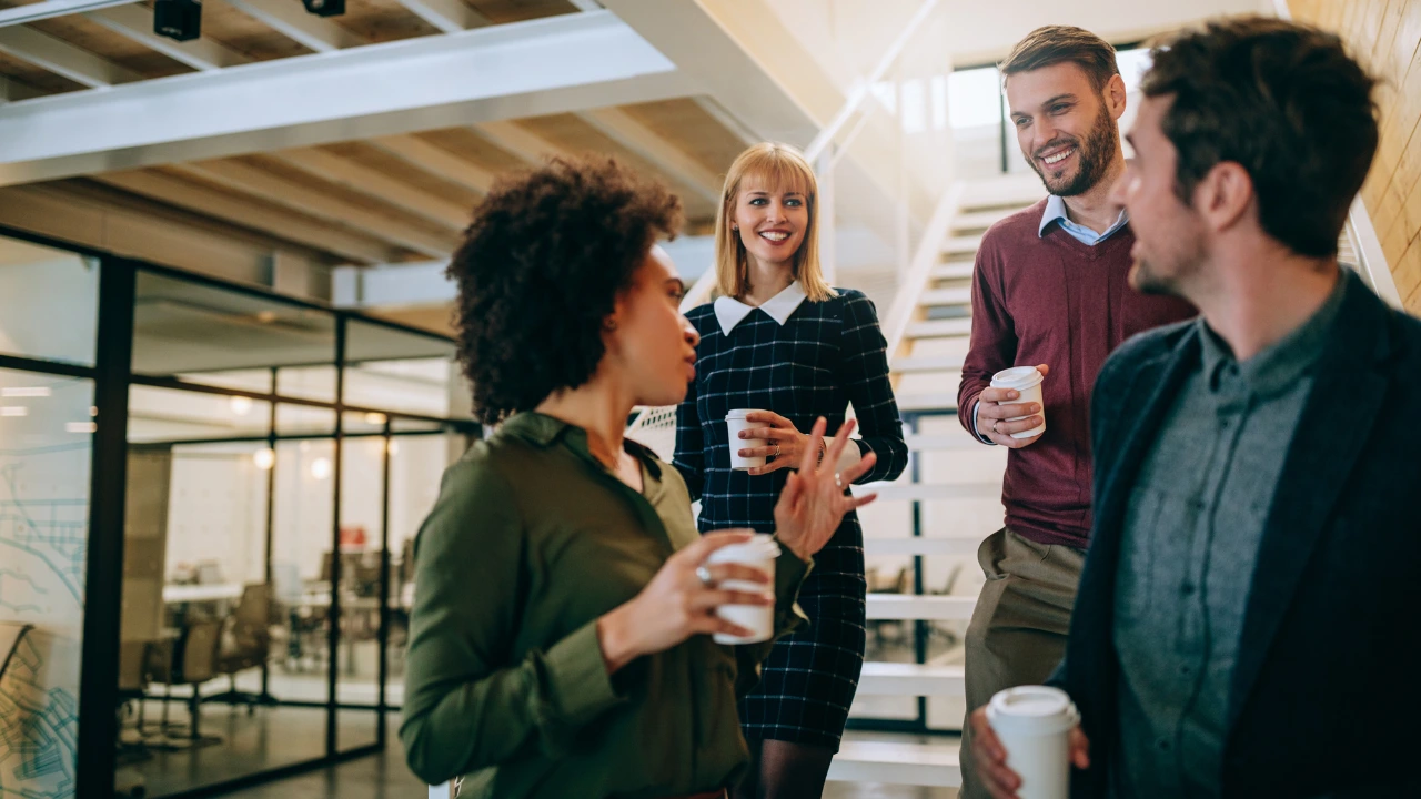 A group of four diverse colleagues chatting and laughing together on a modern office staircase during a breakroom wellness coffee break, holding disposable cups.