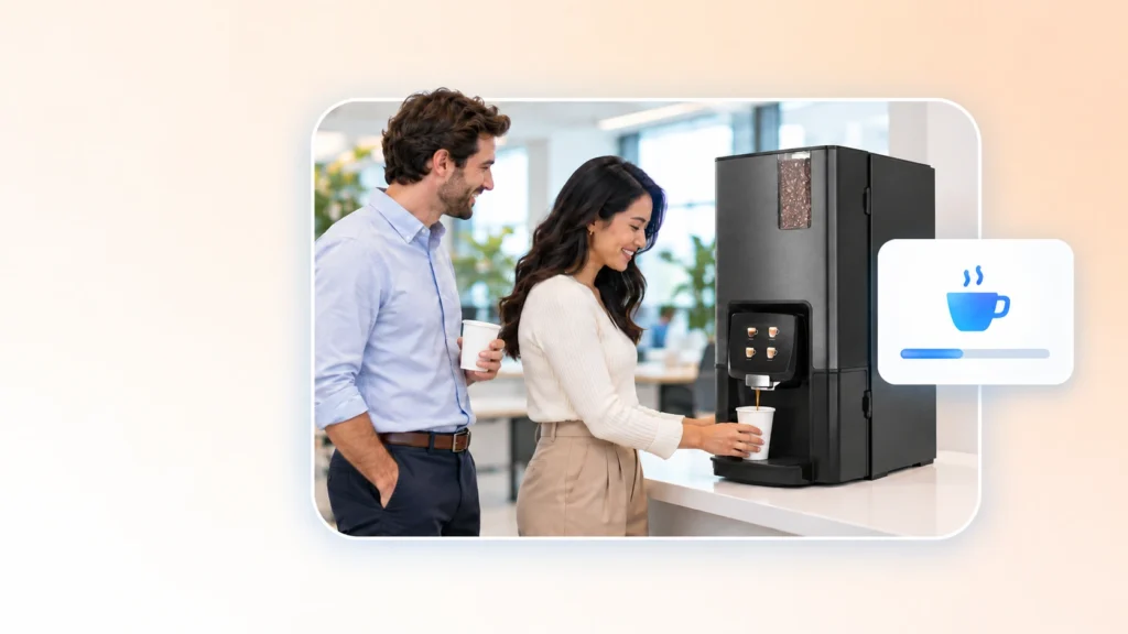 Two coworkers smile as one fills a cup at a workplace coffee machine in a bright office breakroom